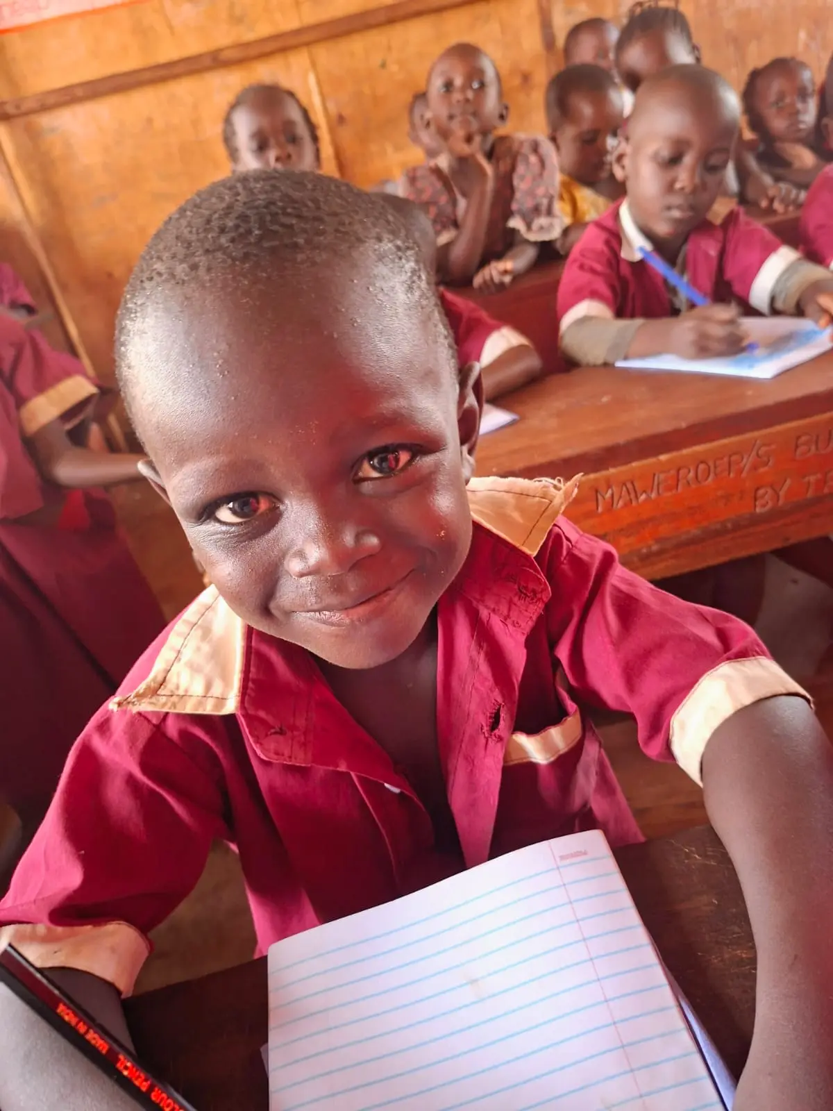 A nursery child in a red uniform smiles while writing in an exercise book at the primary school in Busia