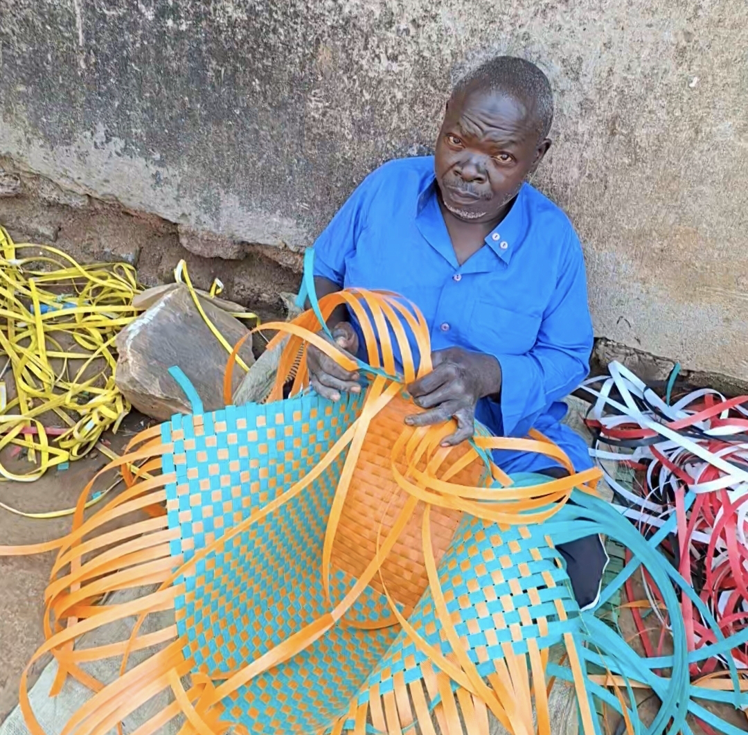 Issa hand-weaving a large orange and turquoise basket at his workspace in Mawero, Busia
