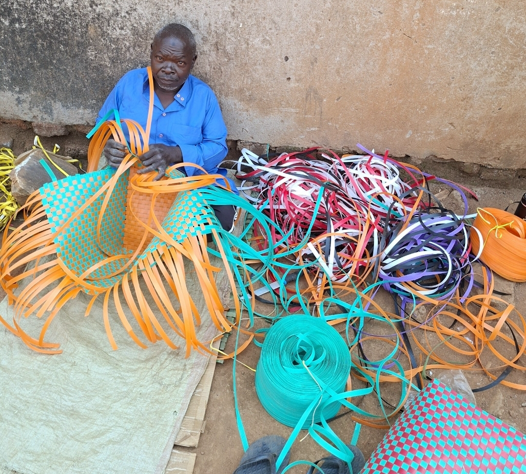 Issa weaving an orange and turquoise basket surrounded by coloured strips bought with his investment in Mawero, Busia